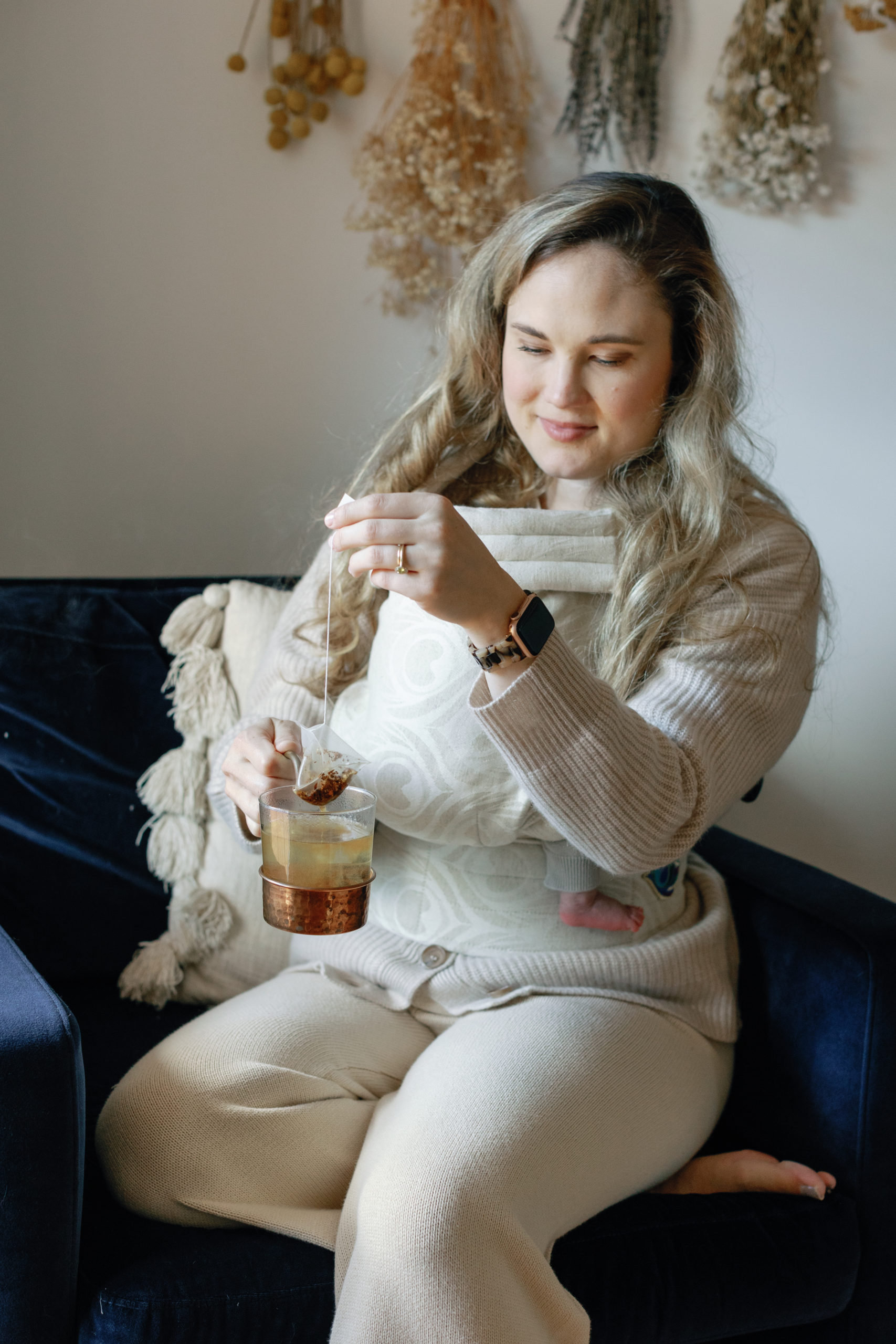 A woman wearing tan wearing her baby in a tan carrier on her front side while holding a tea bag she has dipped in her mug.